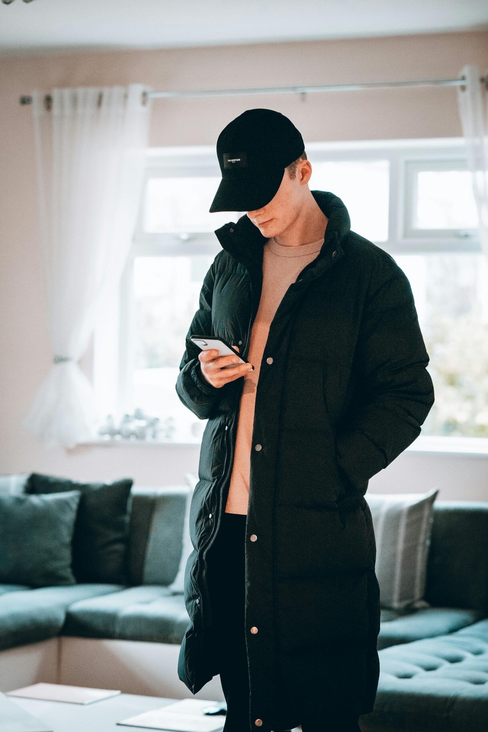 Young man in black winter fashion checks smartphone indoors with window light.