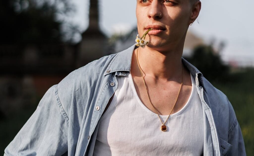 Stylish young man in casual attire poses with flower in mouth outdoors.