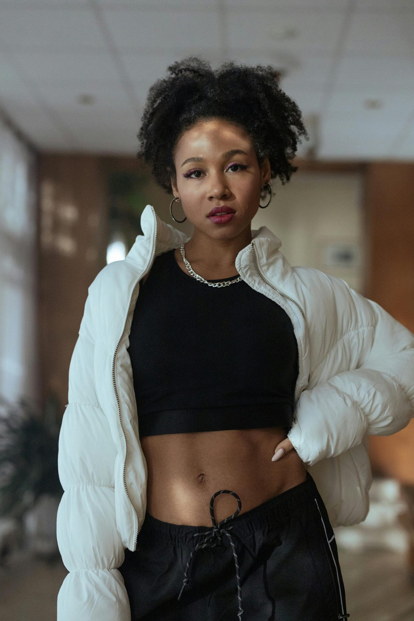 Confident woman with curly hair wearing a crop top and jacket in a modern indoor setting.