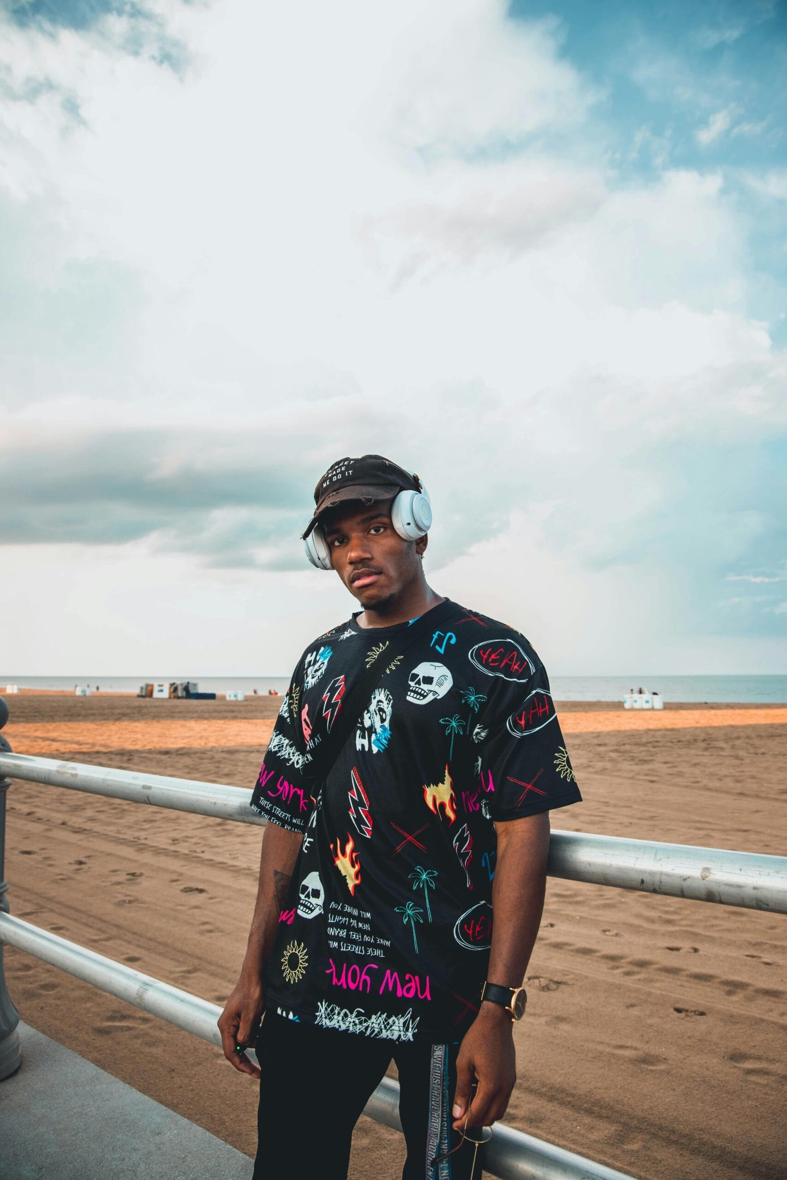 Stylish young man in a graphic T-shirt wearing headphones at the beach with a cloudy sky backdrop.